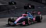 Pink and blue Formula 1 car leads a race on a curved track, with two darker cars in the background.