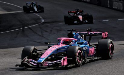 Pink and blue Formula 1 car leads a race on a curved track, with two darker cars in the background.