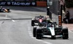 Front row Mercedes F1 car leads a street circuit with other racing cars in pursuit, barrier walls and a TAG Heuer banner in the background.