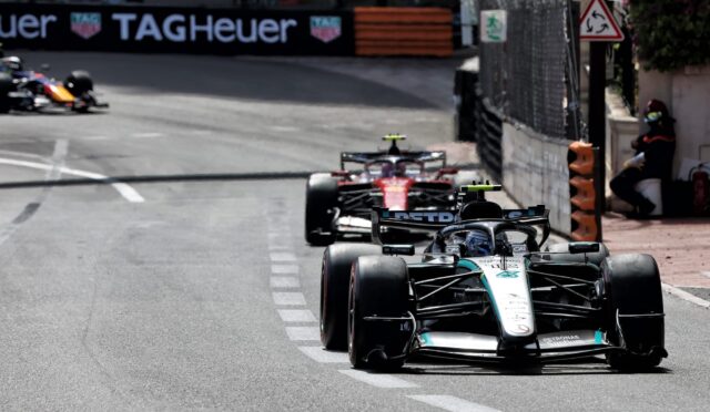 Front row Mercedes F1 car leads a street circuit with other racing cars in pursuit, barrier walls and a TAG Heuer banner in the background.