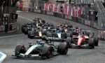 Multiple Formula 1 cars tightly packed as they race along a city street circuit, with barriers and sponsor banners lining the track.