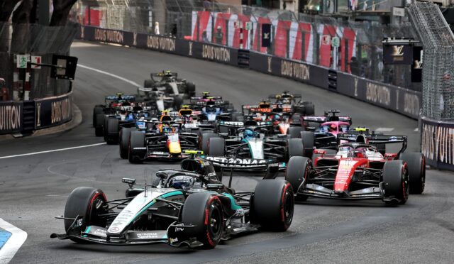 Multiple Formula 1 cars tightly packed as they race along a city street circuit, with barriers and sponsor banners lining the track.