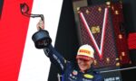 Racing driver in a blue suit lifts a trophy umbrella on a podium, wearing a Pirelli cap, with a red LV-pattern backdrop behind.