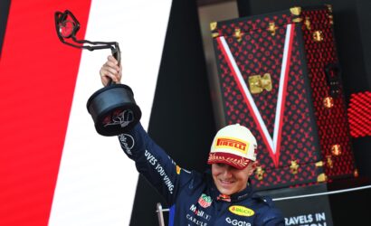 Racing driver in a blue suit lifts a trophy umbrella on a podium, wearing a Pirelli cap, with a red LV-pattern backdrop behind.