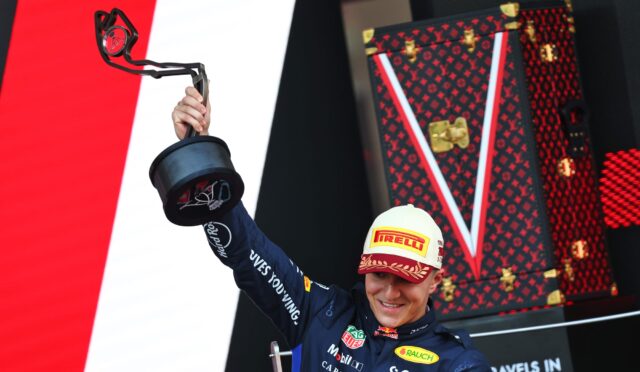 Racing driver in a blue suit lifts a trophy umbrella on a podium, wearing a Pirelli cap, with a red LV-pattern backdrop behind.