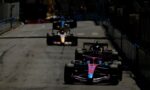 Pit-to-pit street race with multiple Formula 1 cars lined up on a track, foreground pink car leading the pack.