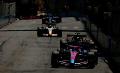 Pit-to-pit street race with multiple Formula 1 cars lined up on a track, foreground pink car leading the pack.