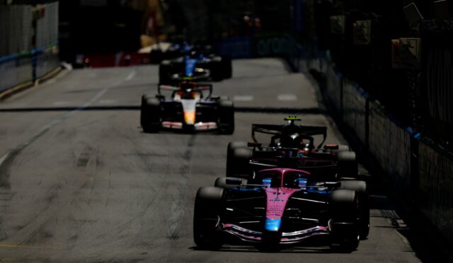 Pit-to-pit street race with multiple Formula 1 cars lined up on a track, foreground pink car leading the pack.