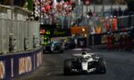 Front-on Formula 1 car races toward camera on a track, with a metal pit wall to the left and a crowded grandstand in the background.