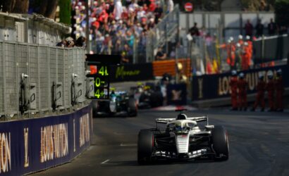 Front-on Formula 1 car races toward camera on a track, with a metal pit wall to the left and a crowded grandstand in the background.