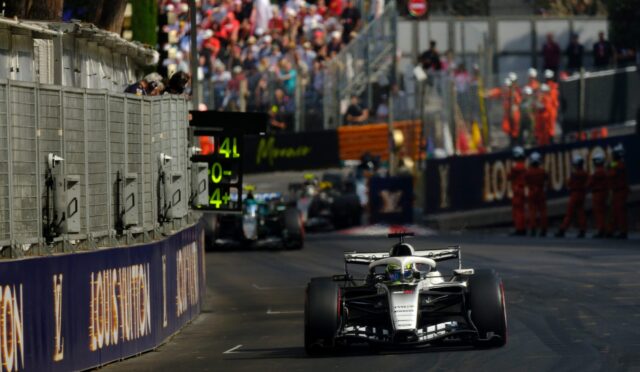 Front-on Formula 1 car races toward camera on a track, with a metal pit wall to the left and a crowded grandstand in the background.