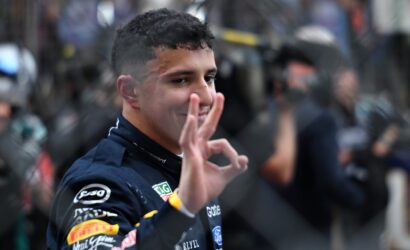 Smiling man in a dark racing suit waves his hand, with a blurred crowd in the background.