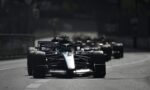 Front Formula 1 car leads a pack on a city street circuit, with other race cars blurred in the background.