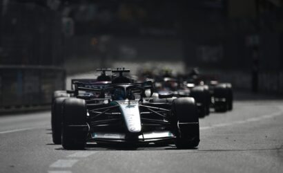 Front Formula 1 car leads a pack on a city street circuit, with other race cars blurred in the background.