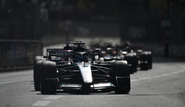 Front Formula 1 car leads a pack on a city street circuit, with other race cars blurred in the background.