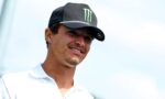 Smiling man wearing a black cap with a green Monster logo outdoors, facing right.