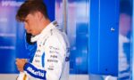 Race car driver in a white and blue sponsor-branded suit standing beside blue lockers, looking down.