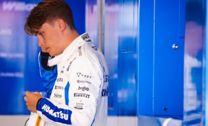 Race car driver in a white and blue sponsor-branded suit standing beside blue lockers, looking down.