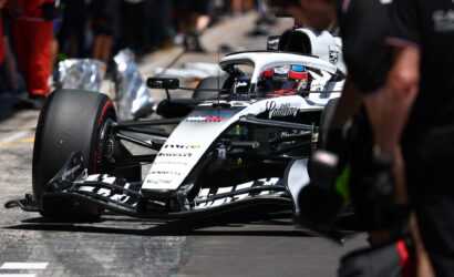 Formula 1 car in the pit lane with a driver in a red helmet, crowd blurred in the background.