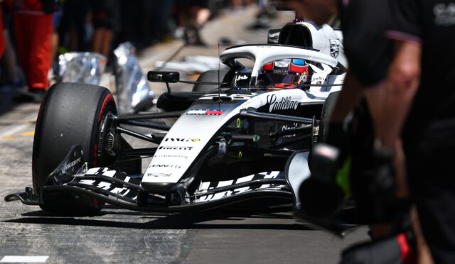 Formula 1 car in the pit lane with a driver in a red helmet, crowd blurred in the background.