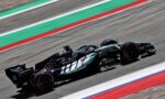Formula 1 car racing at high speed on a track, teal and black livery with sponsor logos visible (Petronas, Microsoft).