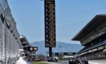 Tall race timing board near pit lane listing driver codes (RUS, PIA, LEC, etc.) and lap times on a sunny day while Formula 1 cars race by nearby.
