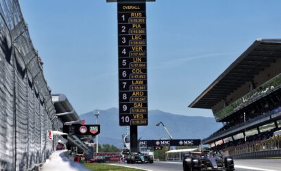Tall race timing board near pit lane listing driver codes (RUS, PIA, LEC, etc.) and lap times on a sunny day while Formula 1 cars race by nearby.