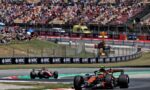 Open-wheel race cars navigate a sharp turn on a racing circuit as a crowded grandstand watches from behind barriers.