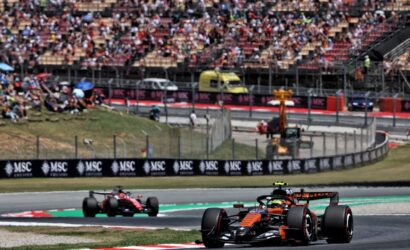 Open-wheel race cars navigate a sharp turn on a racing circuit as a crowded grandstand watches from behind barriers.