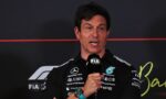Man in a black sponsor-laden shirt speaks into a FIA-branded microphone at a press event.
