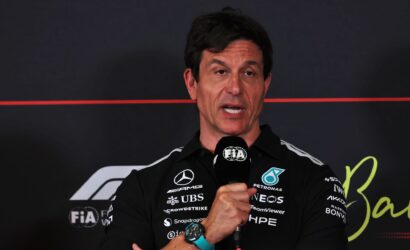 Man in a black sponsor-laden shirt speaks into a FIA-branded microphone at a press event.