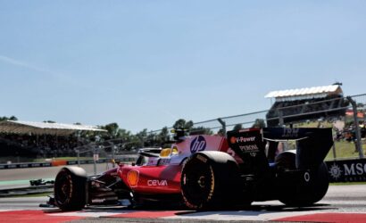 Red and pink Formula 1 car on a sunny race track with grandstands filled with spectators in the background, crossing a red curb point.