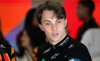 Young male racer in a black Monster Energy racing suit, looking to his right against a red blurred background at a busy event.