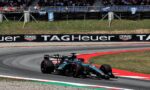 Formula 1 car navigating a left-hand turn on a race track, with spectators in the grandstands and barriers along the course.