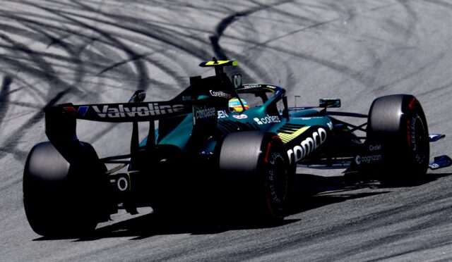 Teal and black Formula 1 car speeding on a racetrack with tire smoke trails behind it.
