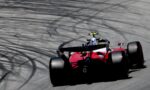 Red Formula-style race car speeding along a track, with dark tire-marked pavement in the background.