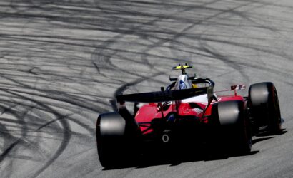 Red Formula-style race car speeding along a track, with dark tire-marked pavement in the background.