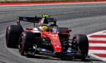 Red Ferrari Formula 1 car on a race track, navigating a turn with close following car and sponsor decals visible on the livery.