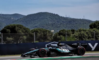 Two Formula 1 cars race on a track, with a chain-link fence and green hills in the background under a blue sky.