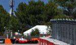 Red Formula 1 race car on a street circuit, driver in white helmet, hugging a barrier with orange safety rails as trees loom in the background.