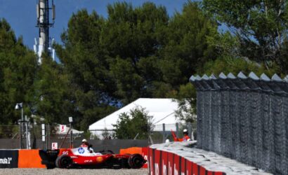Red Formula 1 race car on a street circuit, driver in white helmet, hugging a barrier with orange safety rails as trees loom in the background.