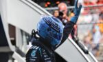 Race car driver in a blue helmet and suit raises a gloved hand in a victory gesture; sponsor logos visible on suit and helmet.
