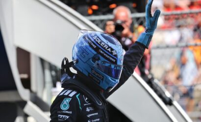 Race car driver in a blue helmet and suit raises a gloved hand in a victory gesture; sponsor logos visible on suit and helmet.