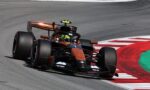 Open-wheel race car in orange and black livery cornering on a race track with red-and-white curb nearby.