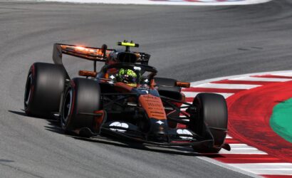 Open-wheel race car in orange and black livery cornering on a race track with red-and-white curb nearby.