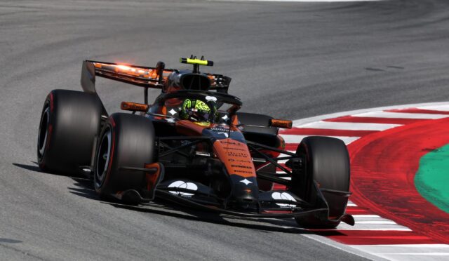 Open-wheel race car in orange and black livery cornering on a race track with red-and-white curb nearby.