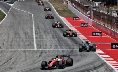 Formula 1 cars racing on a street circuit with red-and-white curbs and barriers along the track.