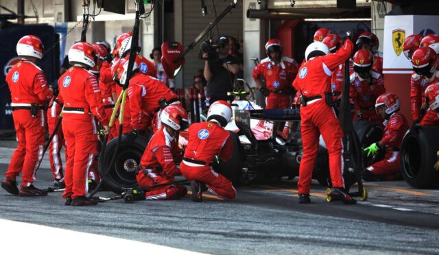 Ferrari pit crew in red uniforms surround a Formula 1 car during a pit stop with tires and equipment nearby