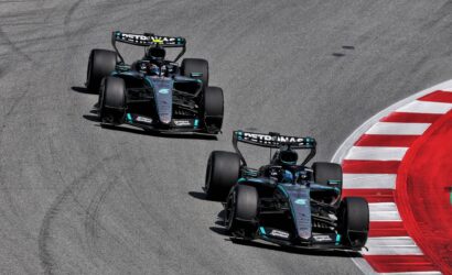 Three Mercedes Formula One cars on a race track, cornering to the left with a red-and-white curb on the right edge.