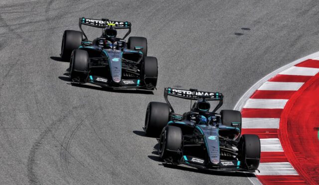 Three Mercedes Formula One cars on a race track, cornering to the left with a red-and-white curb on the right edge.